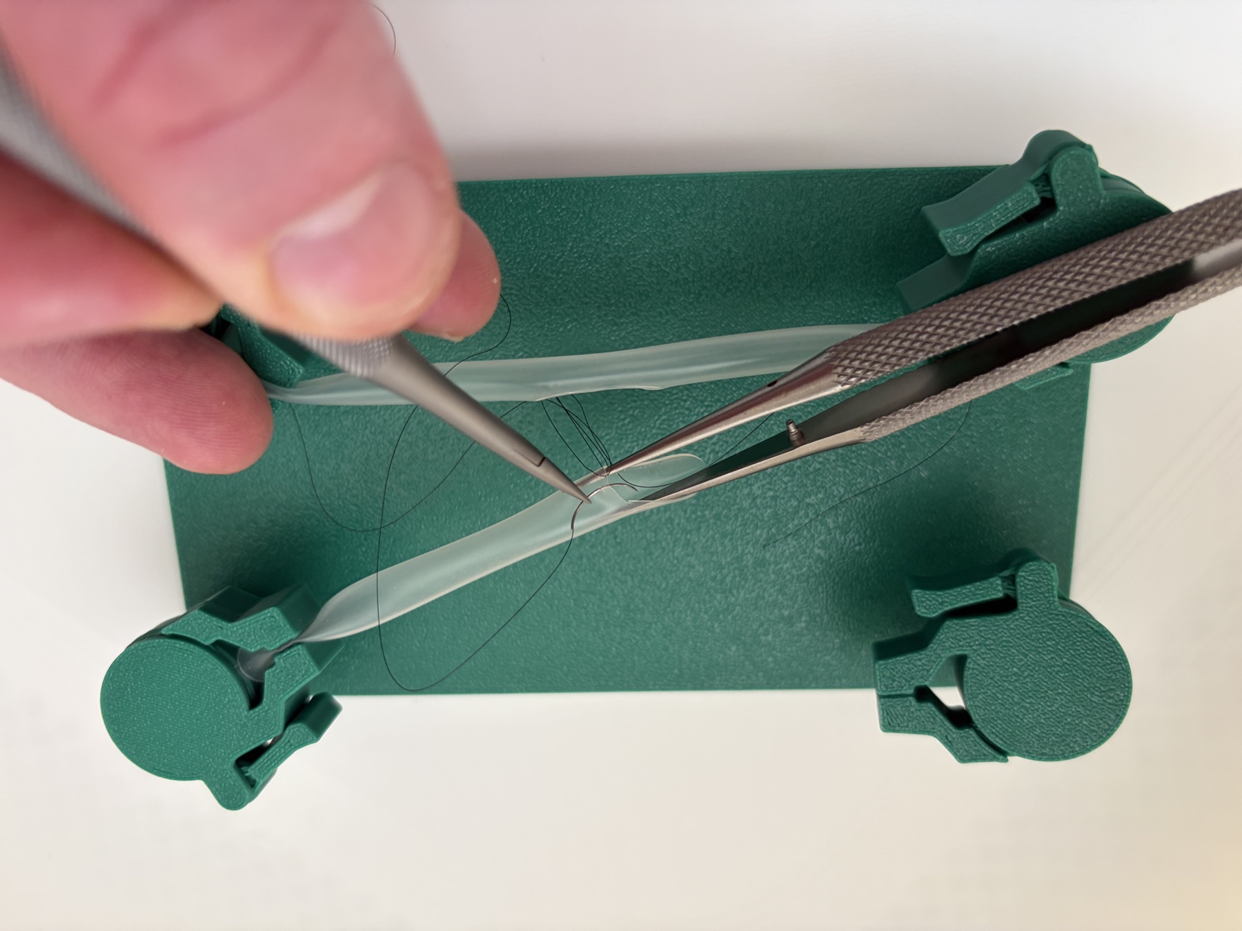 Hands using surgical instruments to practise suturing simulated vessels held by the jig.